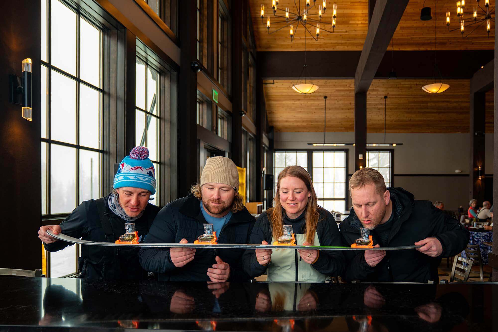 Four people enjoy ski shots indoors at a cozy lodge bar, using a ski to hold their drinks side-by-side.