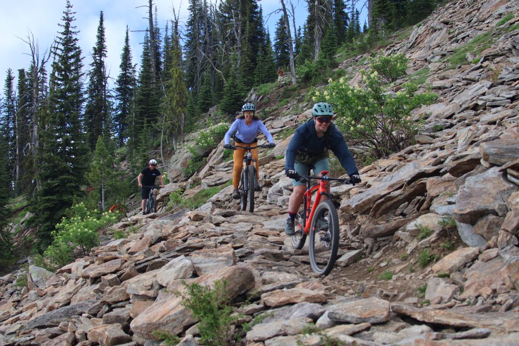 Three people riding mountain bikes along a rocky hillside trail at Brundage Mountain Resort, and in the background, a forest of trees.