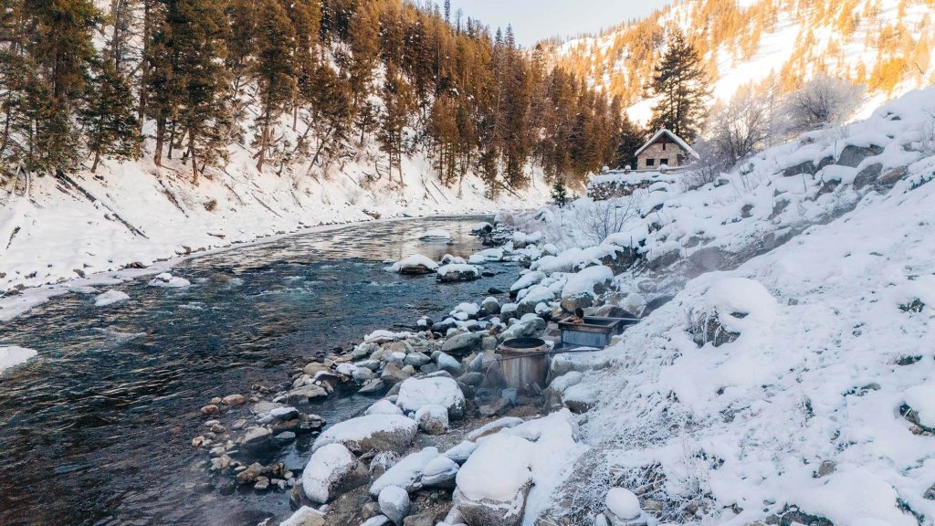 A person relaxes in a steaming tub at Sunbeam Hot Springs along the Salmon River, surrounded by snow-covered rocks and a rustic cabin near Stanley, Idaho