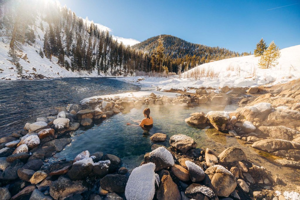 Woman soaking in a riverside hot spring surrounded by snowy hills and pine trees