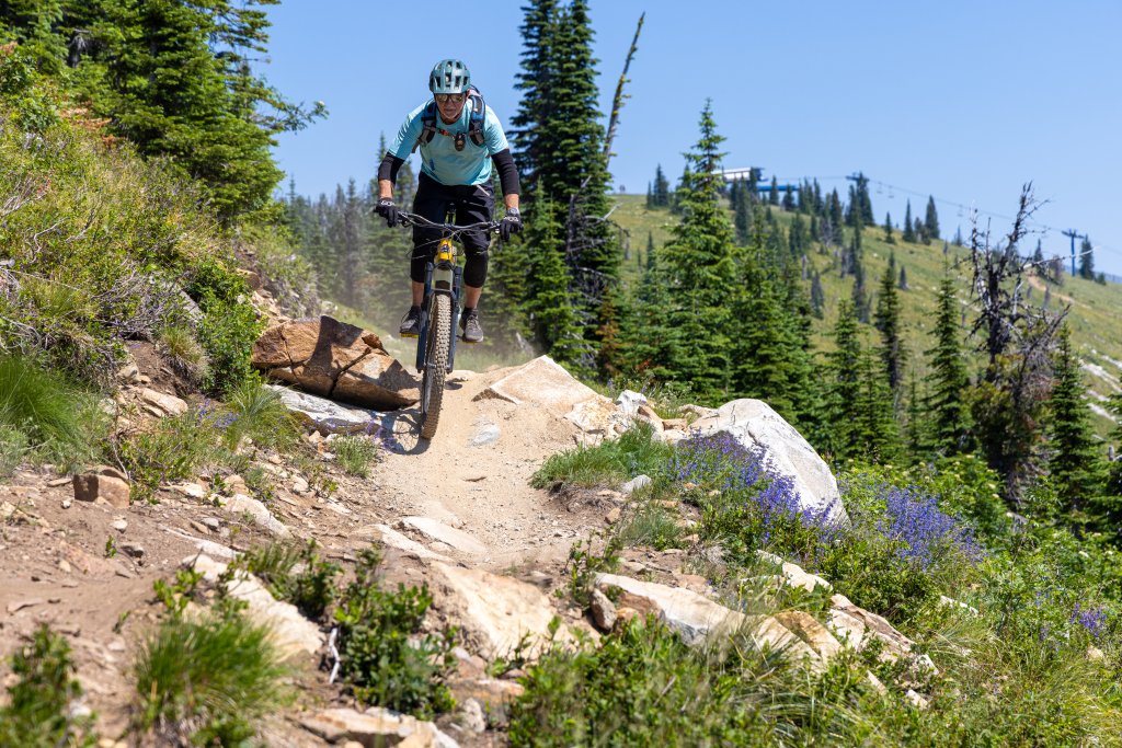 A person riding a mountain bike down a rocky hillside trail at Schweitzer, and in the background, a forest of tall trees.