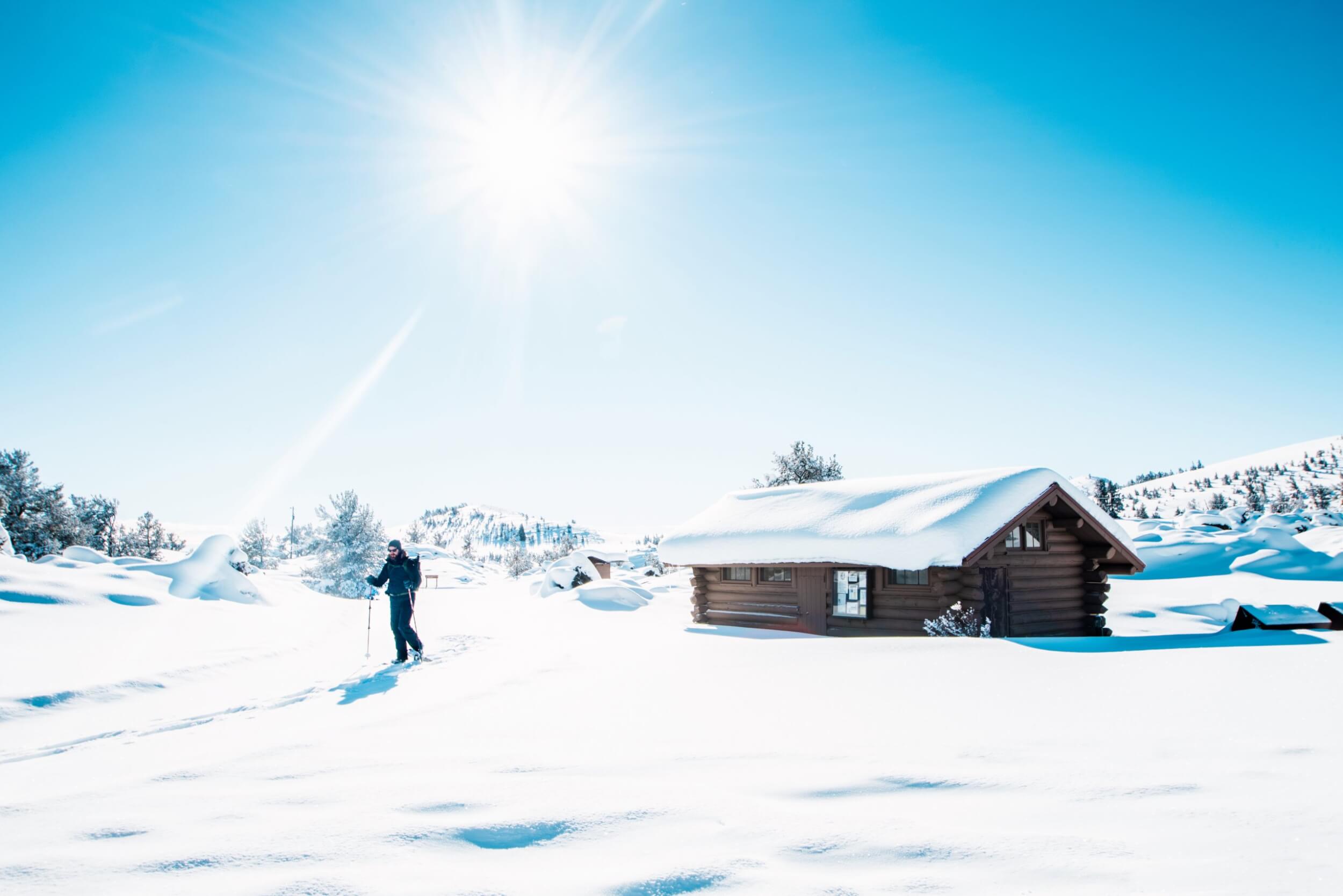 A person cross-country skis on a sunny day next to a snow-covered log cabin.