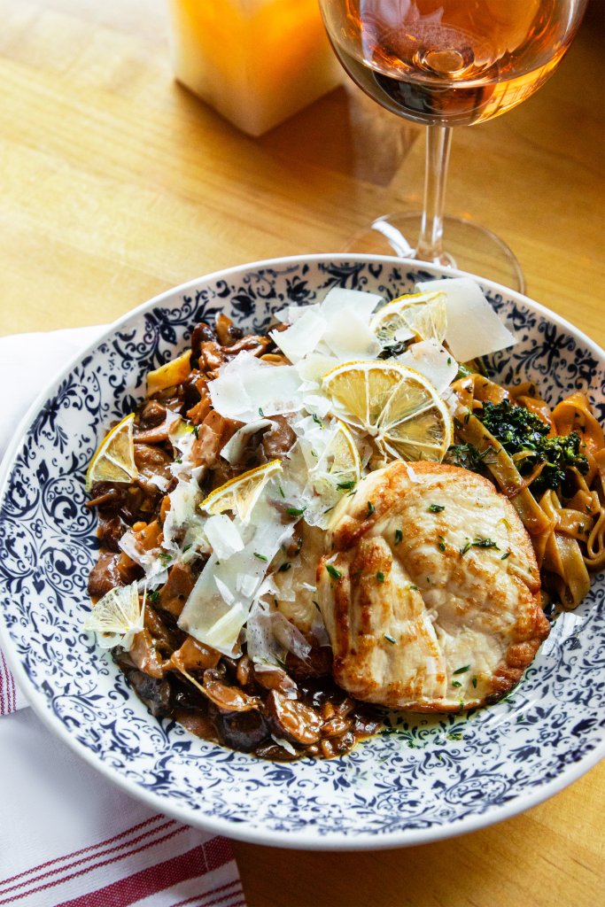 A dish of pasta covered in sauce and grated parmesan beside a glass of wine on a table at ÁLAVITA.