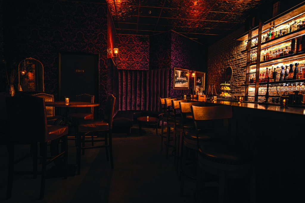 A dimly lit view of the Gatsby bar in Boise, with a table, bar chairs, glowing bottles and Prohibition-era vibes.
