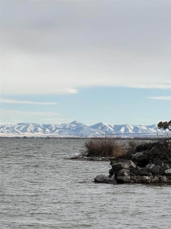 On a winter day, a gray lake is bordered by a rocky shoreline on one side with snowcovered mountains rising in the distance.