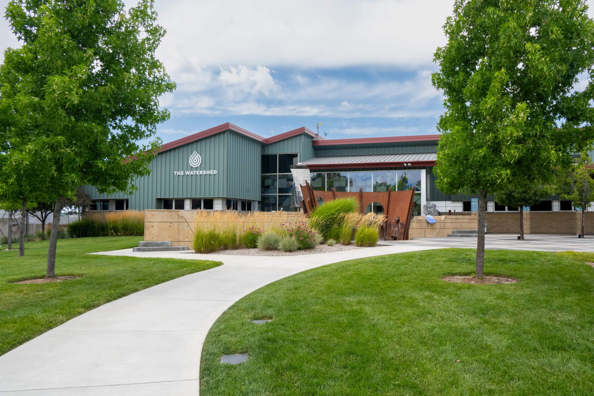 The sage green building sits in the background with a sidewalk and green space leading up to a the entrance featuring native wildflowers and tall grasses.