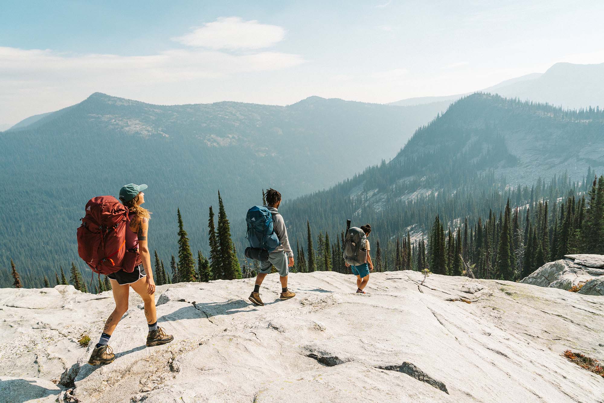 Three hikers with backpacks hike into the Selkirk Mountains, overlooking forest and peaks between Harrison Lake and Beehive Lake near Sandpoint, Idaho.