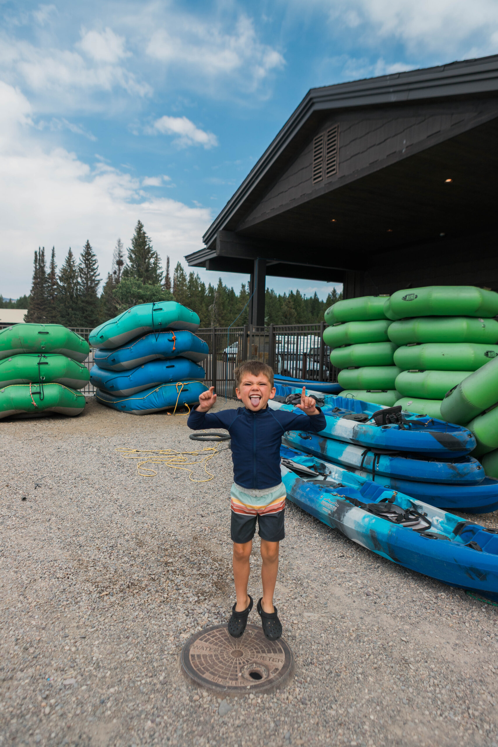 A young boy in a blue swimsuit and swim trunks stands in the middle of stacked rafts, float tubes and kayaks