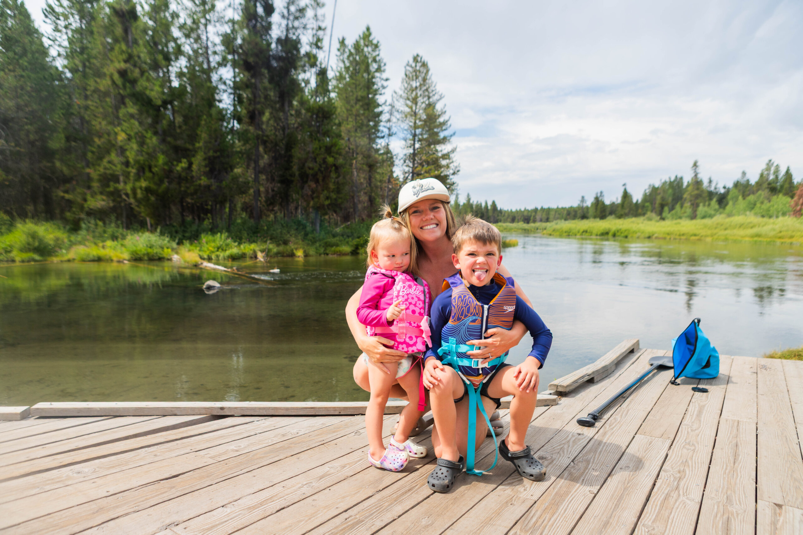 A blonde woman squats down with a young girl in a pink life jacket and a young boy with a blue life jacket on, standing on a dock with a calm river in the background.