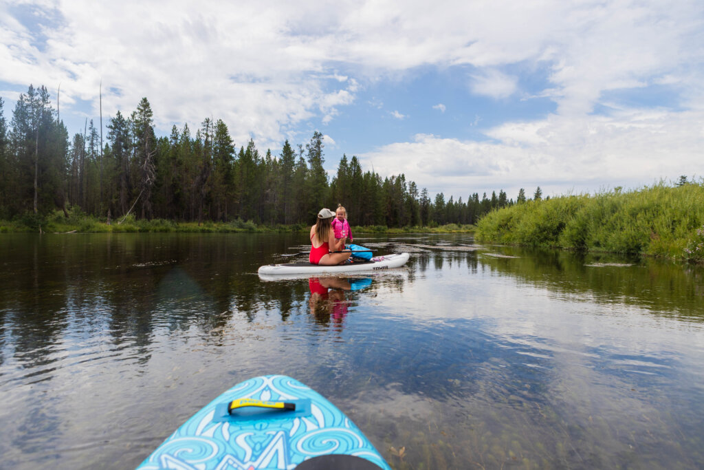 a woman in a red swimsuit and a toddler in a pink swimsuit and life jacket sit on a standup paddleboard on a smooth river
