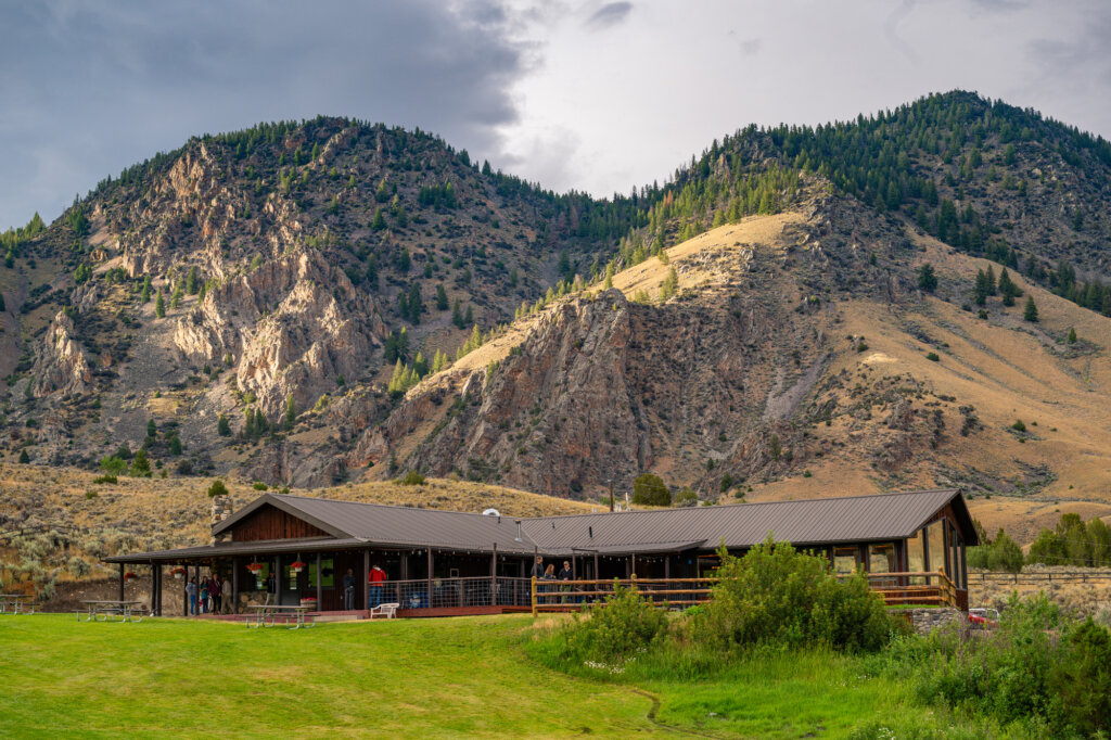 The sun shines on rugged and jagged mountains that sit behind a rustic, wooden lodge at Twin Peaks Ranch State Park in Salmon, Idaho.
