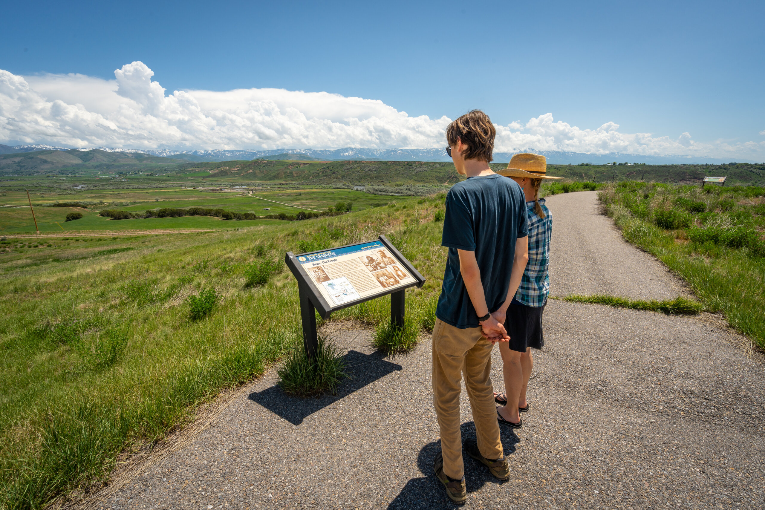 Two visitors read a historical interpretive sign on a paved overlook trail, with a green valley and snow-capped mountains in the distance.