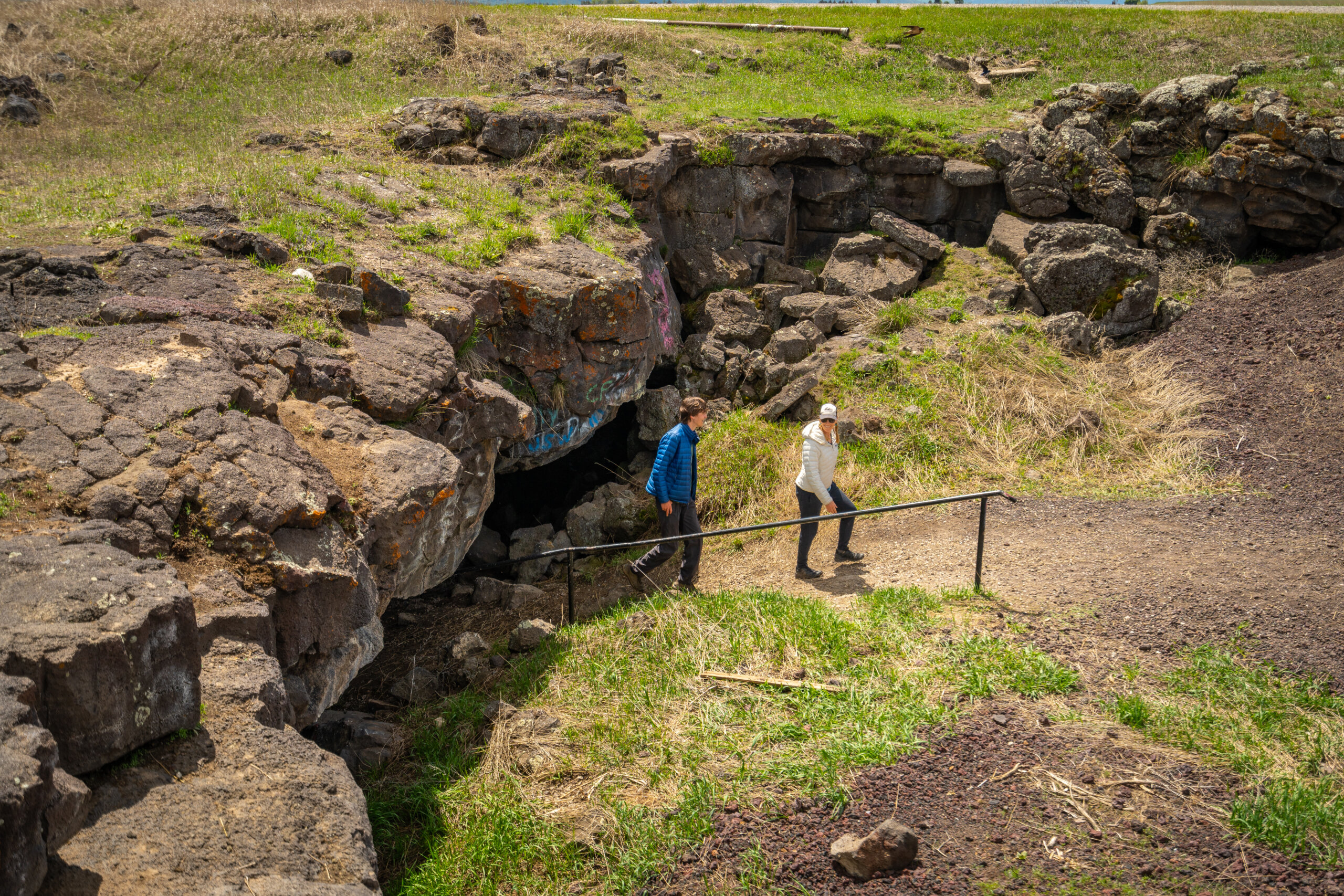 Two hikers in jackets walk down a path toward a dark cave opening in a rocky lava outcropping, with a metal handrail alongside the trail.