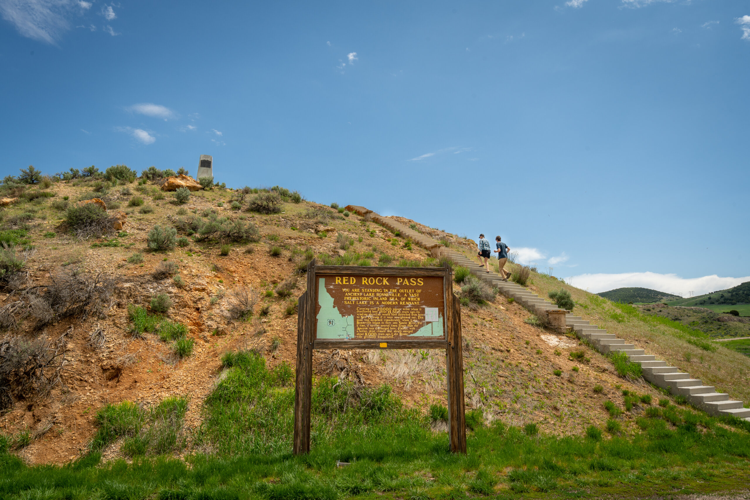 Two hikers climb a concrete staircase up a grassy hillside toward a stone monument, with a wooden interpretive sign reading "Red Rock Pass" in the foreground.