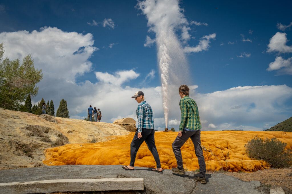 A tall geyser erupts against a blue sky as two visitors walk past an orange mineral-streaked rock formation, with other onlookers watching in the background on the Pioneer Historic Byway.