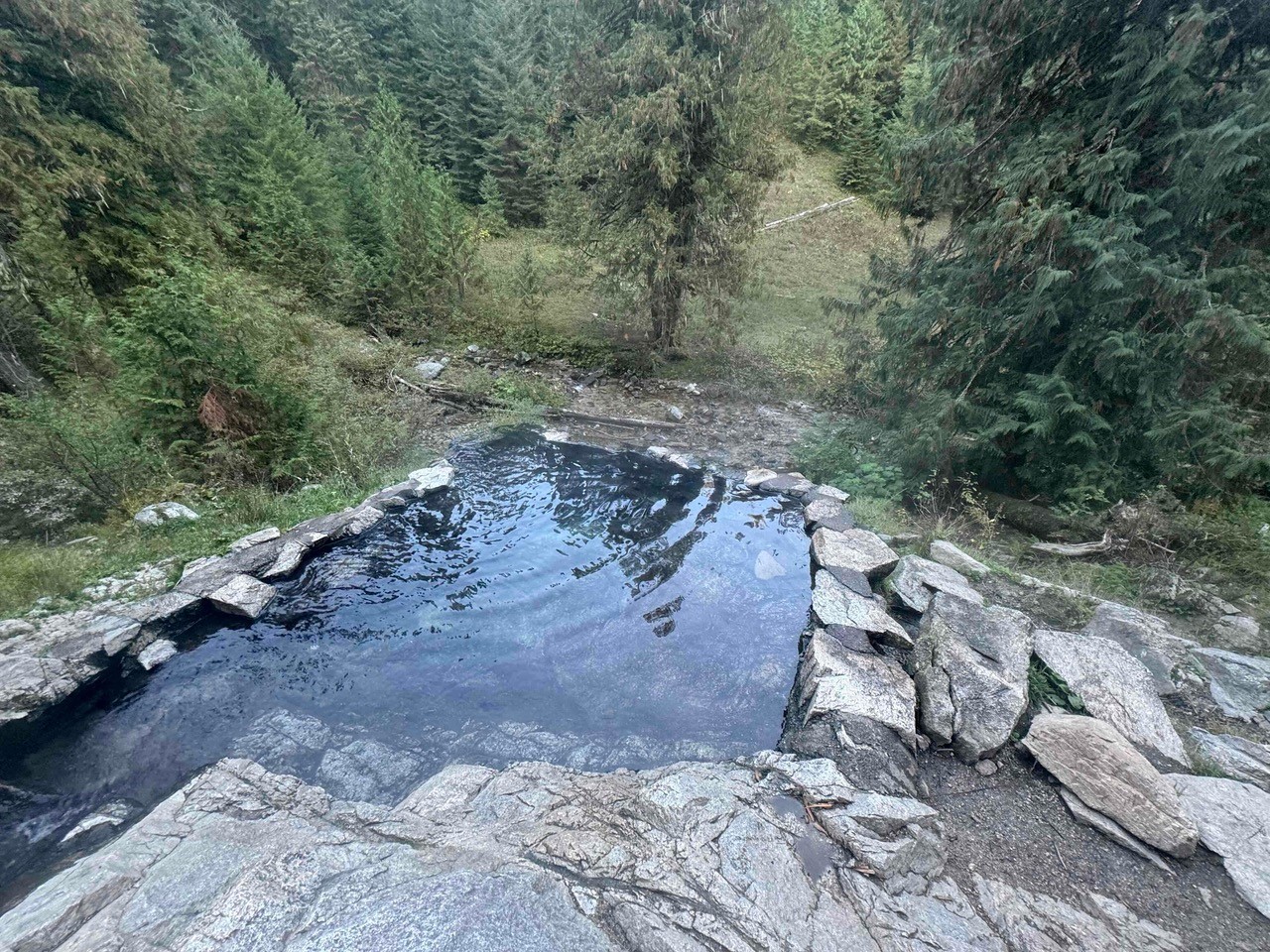A single rock-walled pool holds still, dark water at Weir Creek Hot Springs, tucked into a forest clearing along the Lochsa River corridor.
