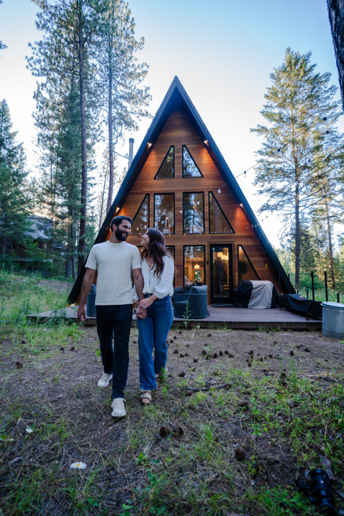 A couple holds hands walking away from a wooden A-frame cabin in a pine forest at dusk, string lights glowing along the eaves and a covered grill visible on the deck behind them.