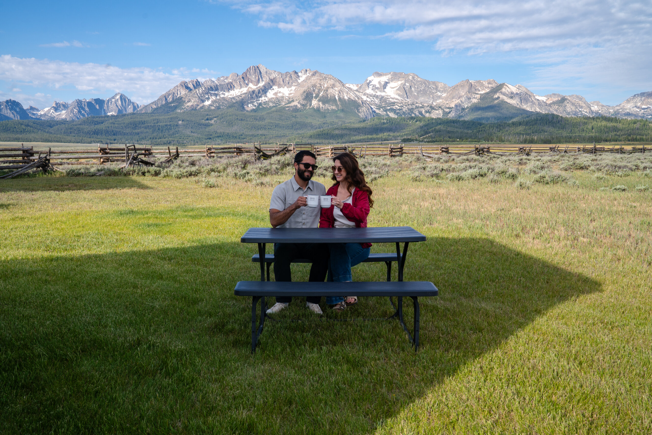 A couple raises coffee mugs in a toast at a picnic table in a green meadow, with a split-rail fence and the jagged, snow-dusted Sawtooth Mountains rising behind them.