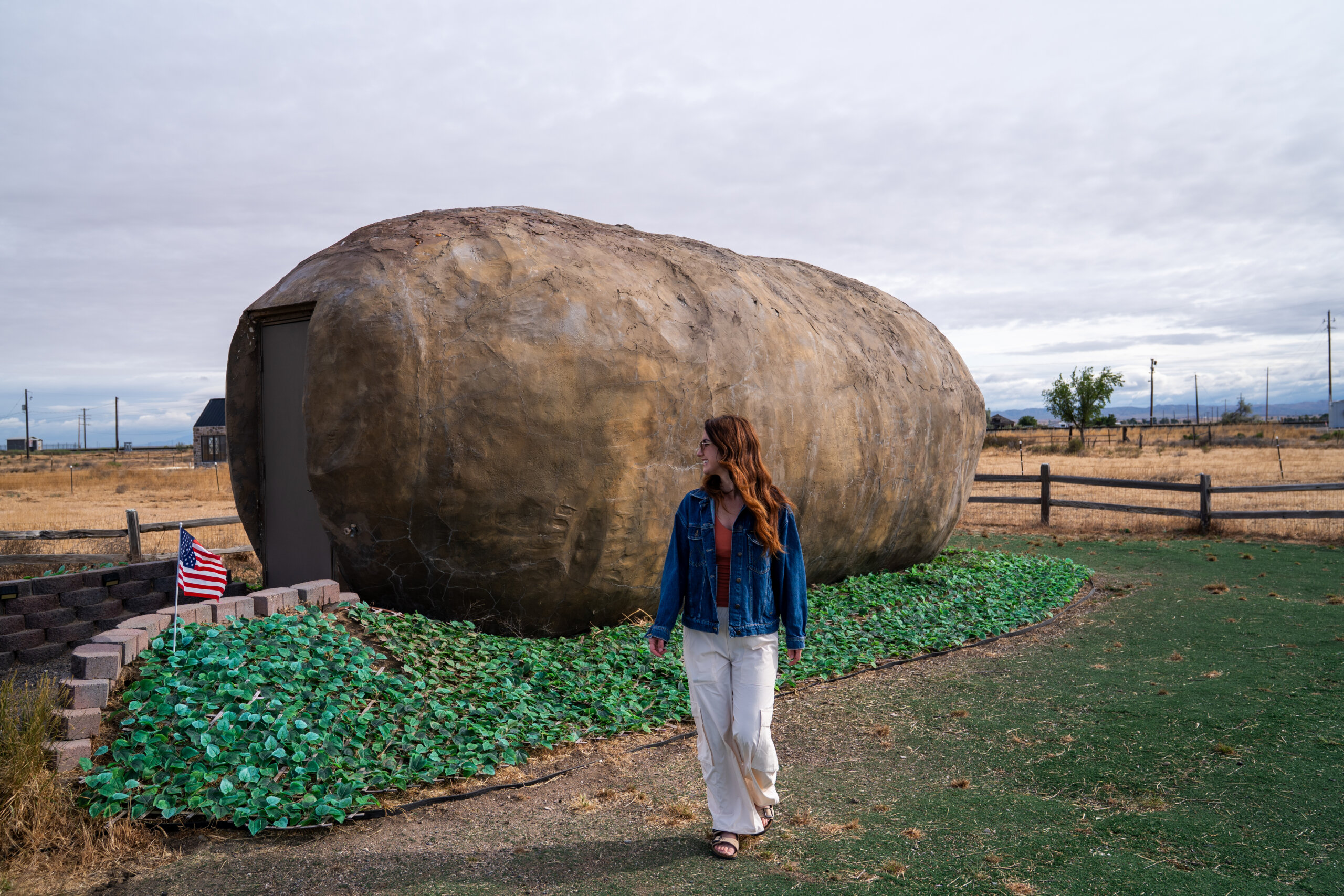 A woman in a denim jacket walks past the Idaho Potato Hotel, a giant potato-shaped building with a door built into its side, set on a grassy lawn under an overcast sky.
