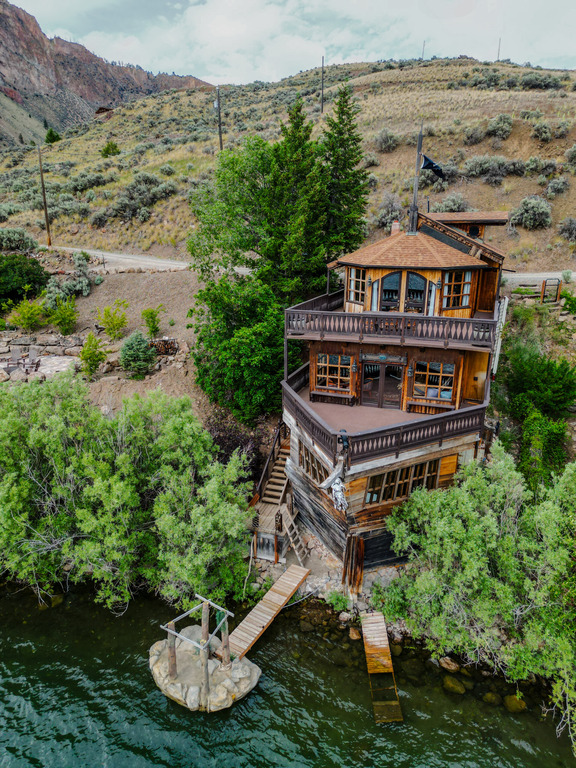 An aerial view of a rustic three-story wooden lodge resembling a wooden ship built into a hillside above, with balconies overlooking the water and a small dock leading down to it.