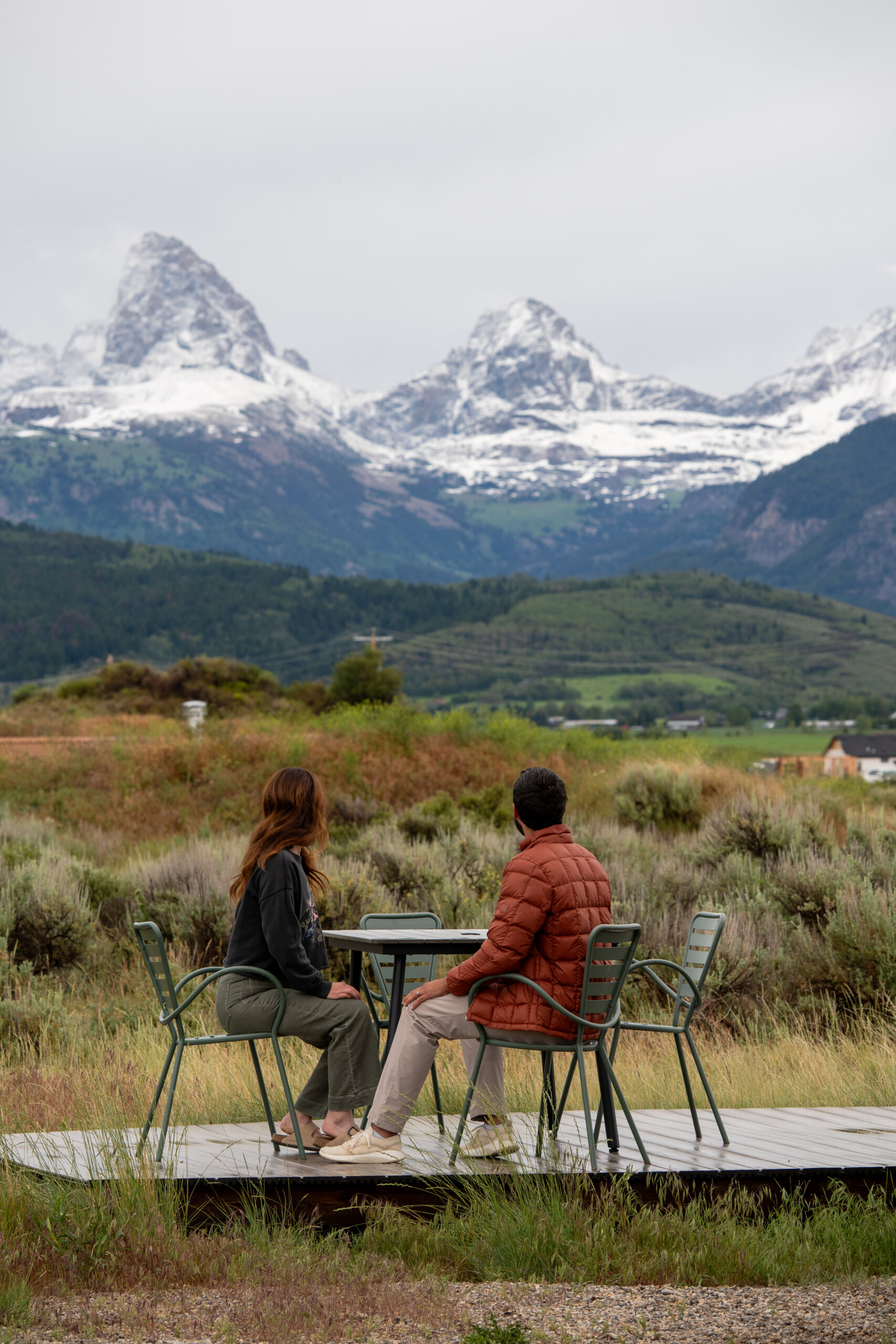 Two people sit at an outdoor table on a wooden platform in a sagebrush field, facing the snow-capped peaks of the Teton mountain range under cloudy skies.