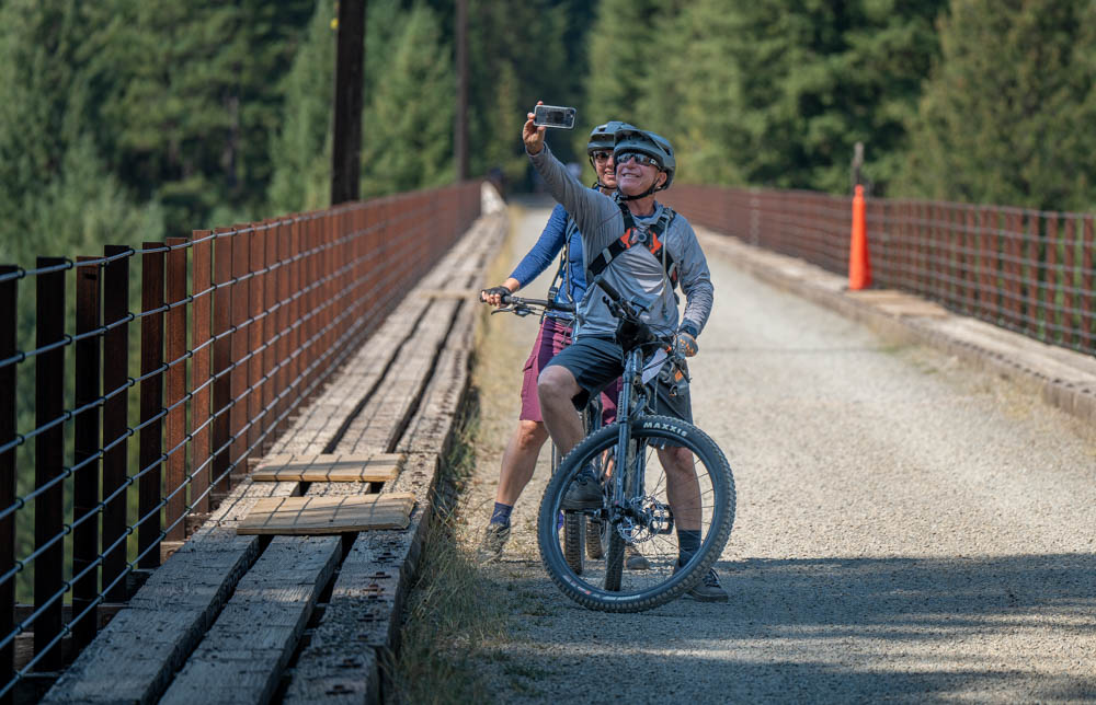 Two bicyclists stand close together on the gravel path of the Route of the Hiawatha while holding a cell phone to take a selfie