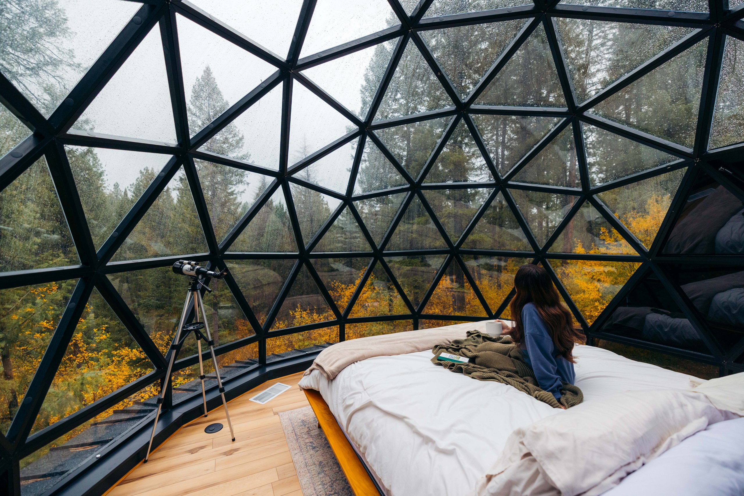 A woman relaxes on a bed inside a geodesic glass dome, with a telescope set up beside her overlooking a forest of fall foliage.