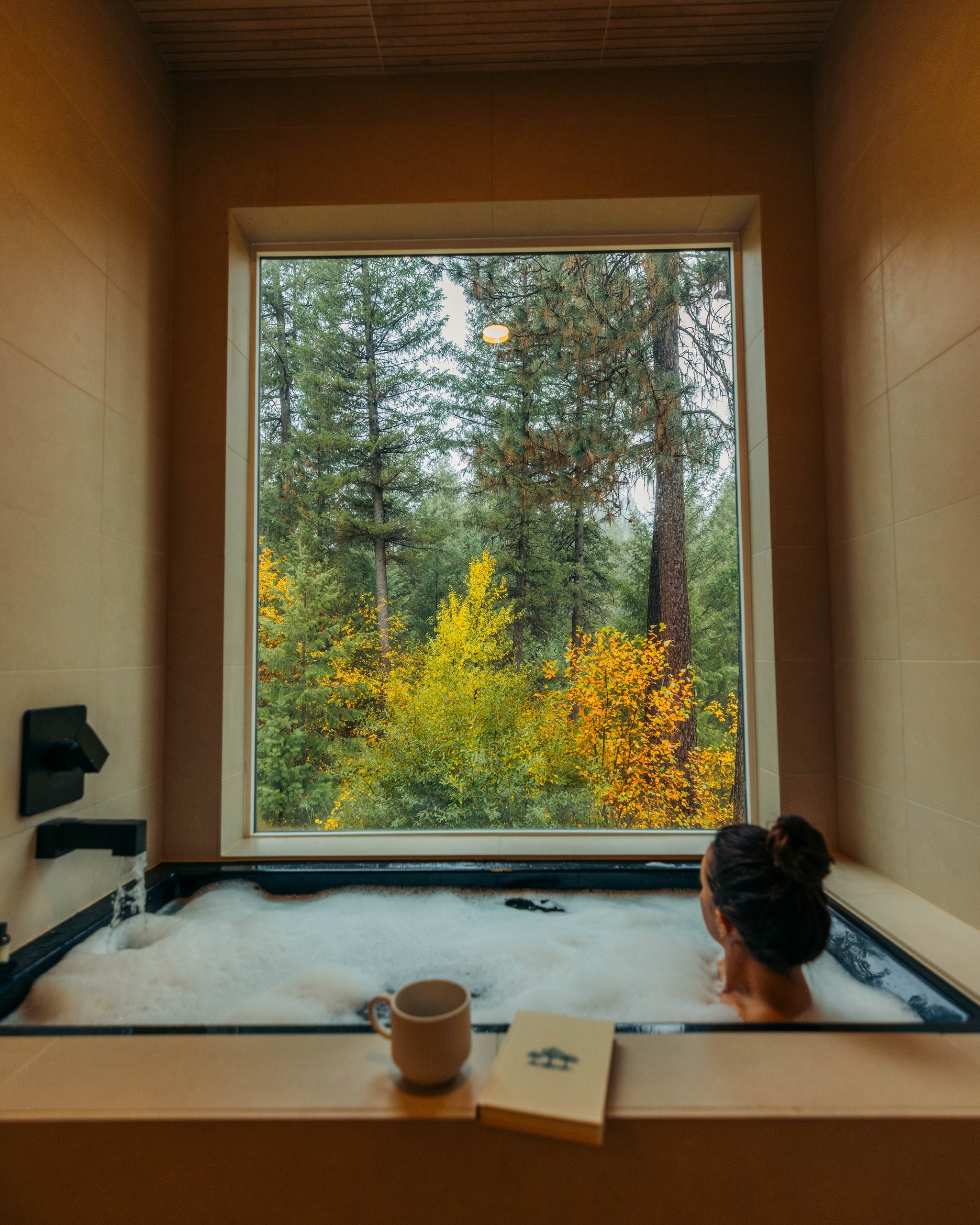 A woman soaks in a bubble-filled bathtub facing a large window overlooking fall foliage, with a mug and book resting on the ledge.