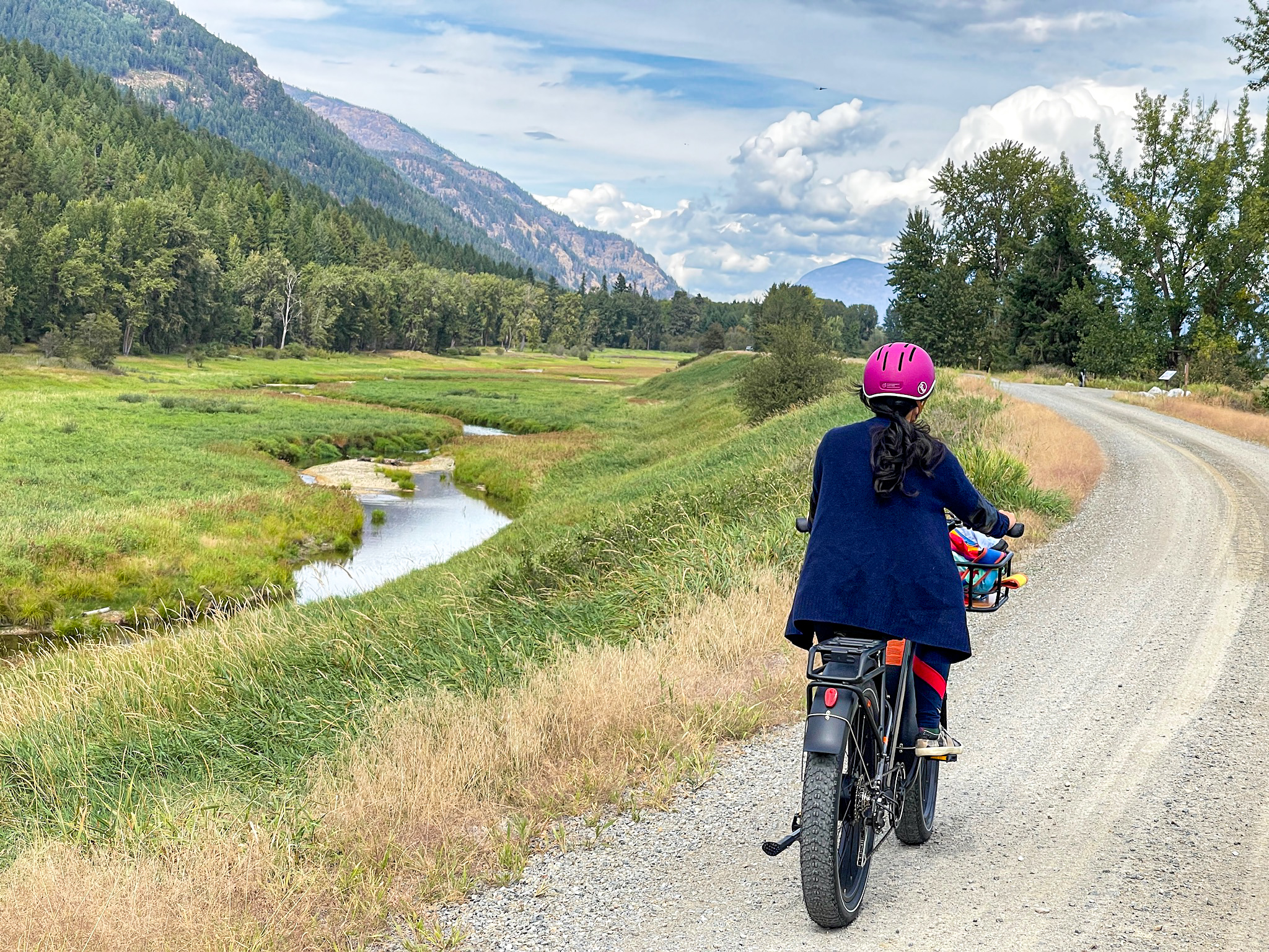 A cyclist in a pink helmet rides a gravel road running beside Myrtle Creek, with a wide grassy valley and forested mountains stretching into the distance under a partly cloudy sky.