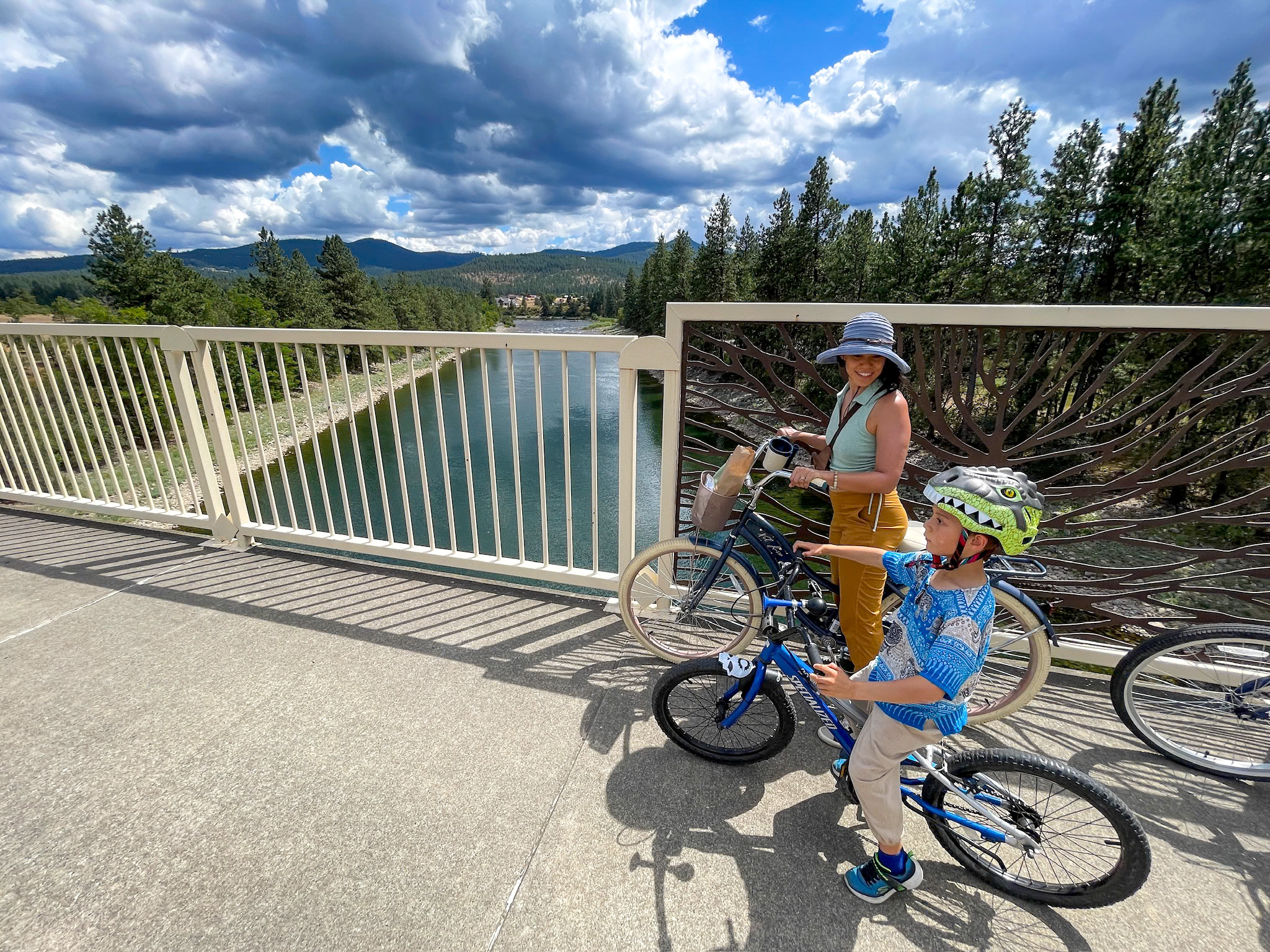 A woman in a sun hat and a boy wearing a dinosaur bike helmet stand with their bicycles on Gateway Bridge, looking out over the Spokane River toward pine-covered hills and a dramatic cloudy sky.