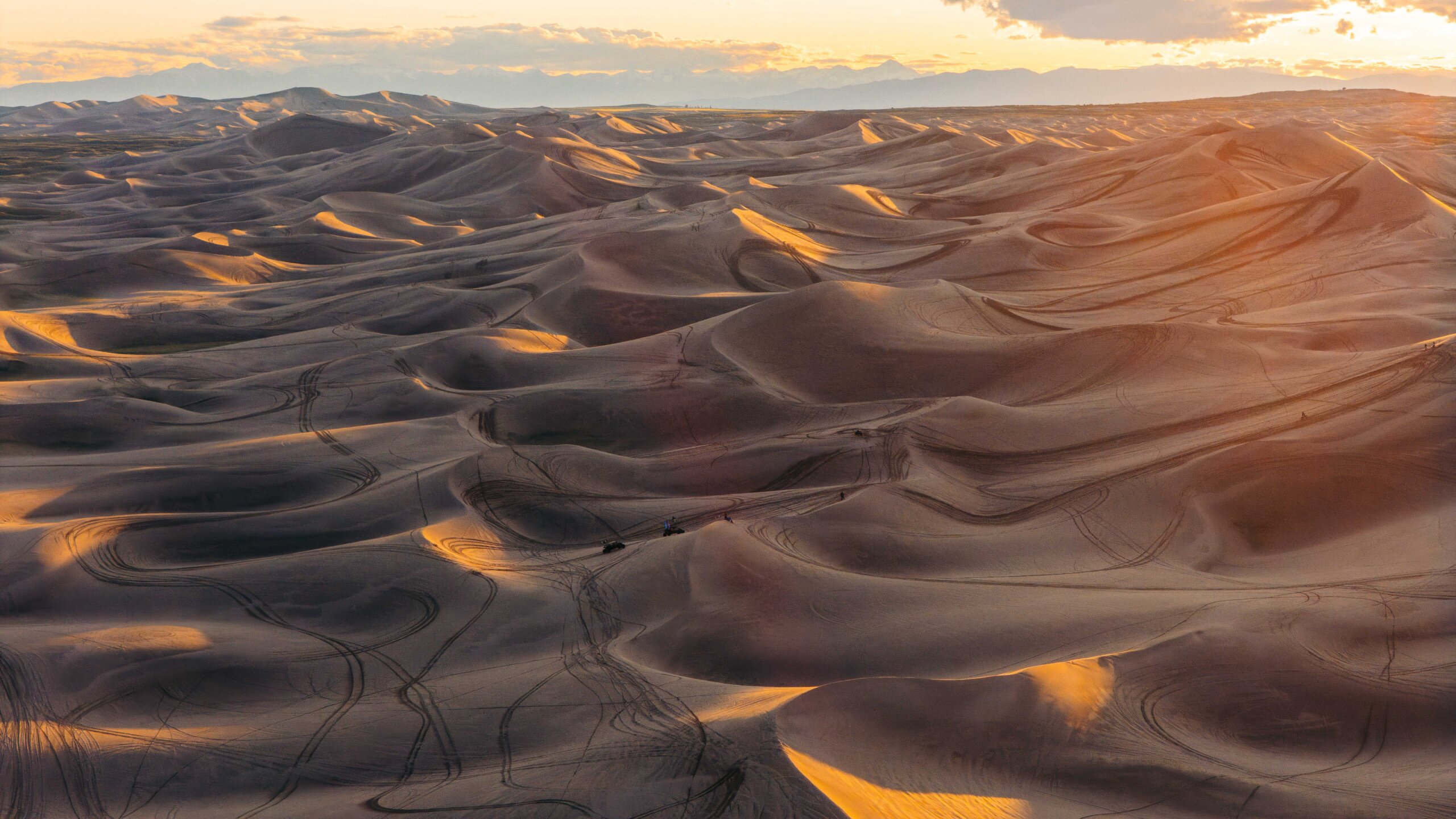 A wide view of rolling St. Anthony sand dunes at golden hour. Low sunlight catches the crests in warm orange and gold, and the troughs between them fall into deep shadow. The dunes stretch in waves to a hazy horizon under a pale sky.
