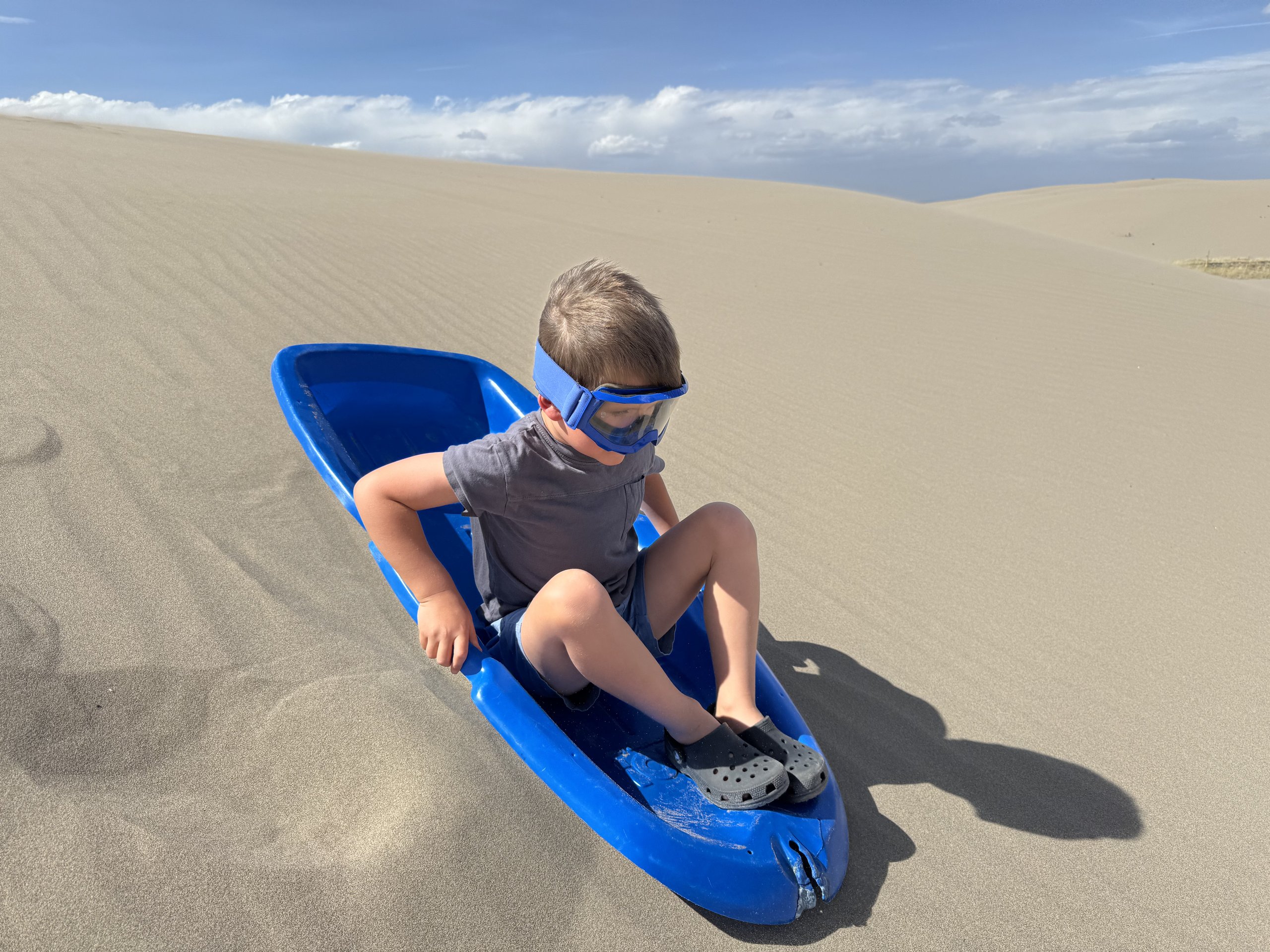 boy wearing sunglasses sits in a blue plastic sled at the top of a sand dune at St. Anthony Sand Dunes, with rolling sand hills and a blue sky in the background.
