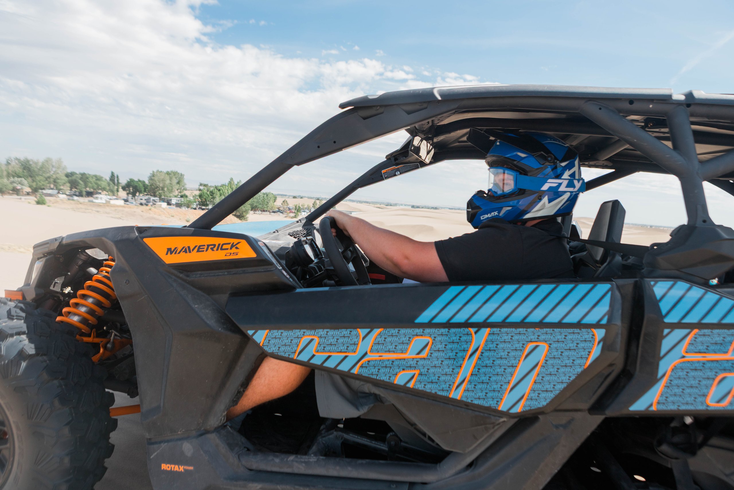 A person drives an off-road UTV across the sand at St. Anthony Sand Dunes, with dune ridges and trees visible in the background.