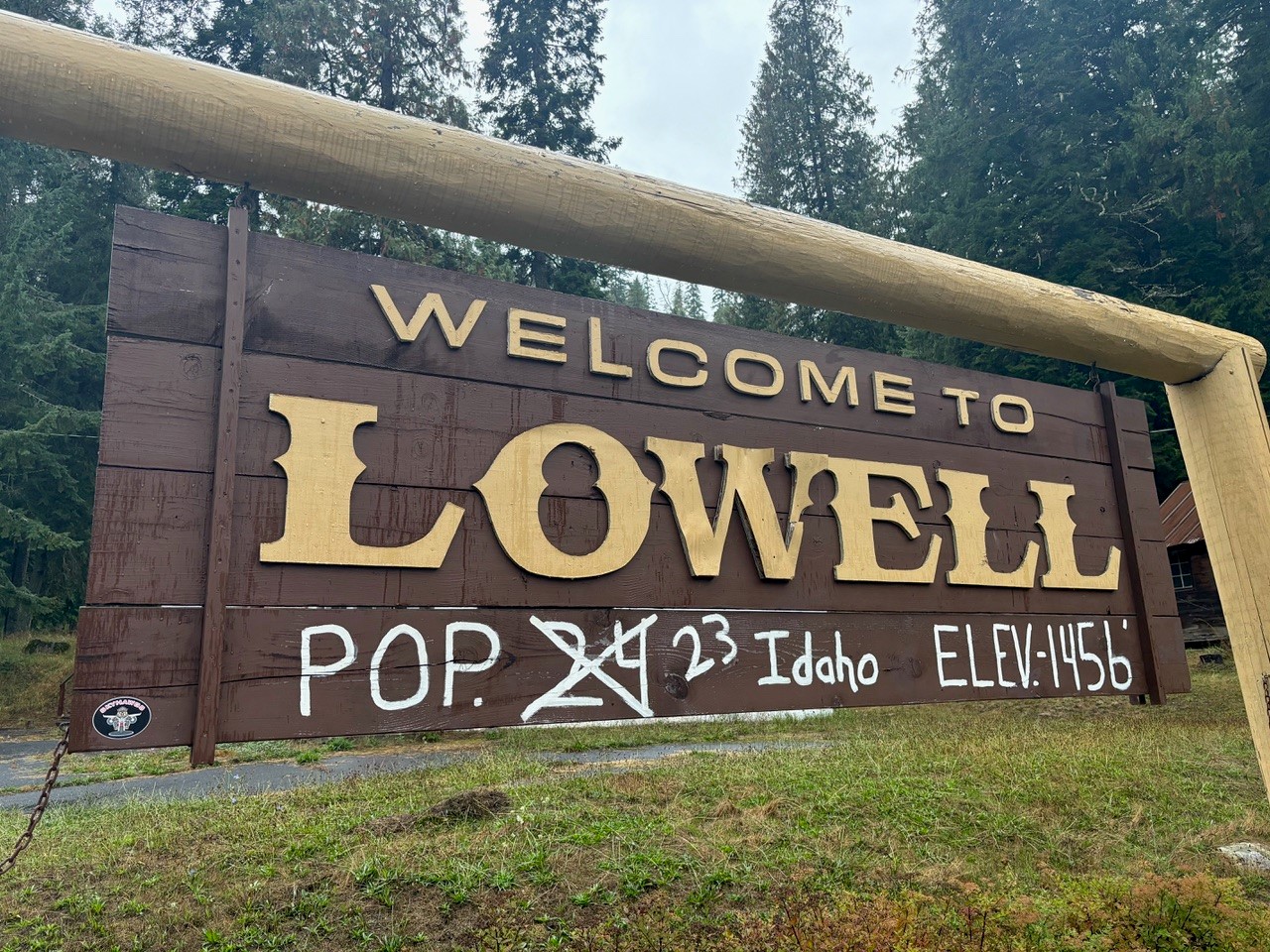 Hand-painted wooden welcome sign for Lowell, Idaho, listing a population of 23 and an elevation of 1,456 feet, framed by dense forest