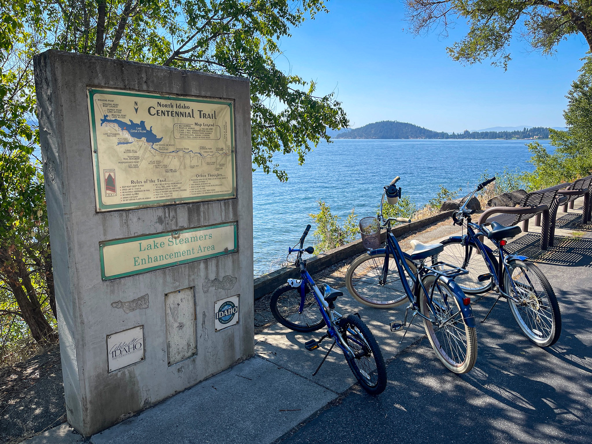 A concrete trail marker with a map of the North Idaho Centennial Trail and a "Lake Steamers Enhancement Area" plaque, standing beside a paved lakeside path where three bicycles are parked. Lake Coeur d'Alene spreads out under a blue sky, with a forested island visible in the distance and a park bench along the shoreline.