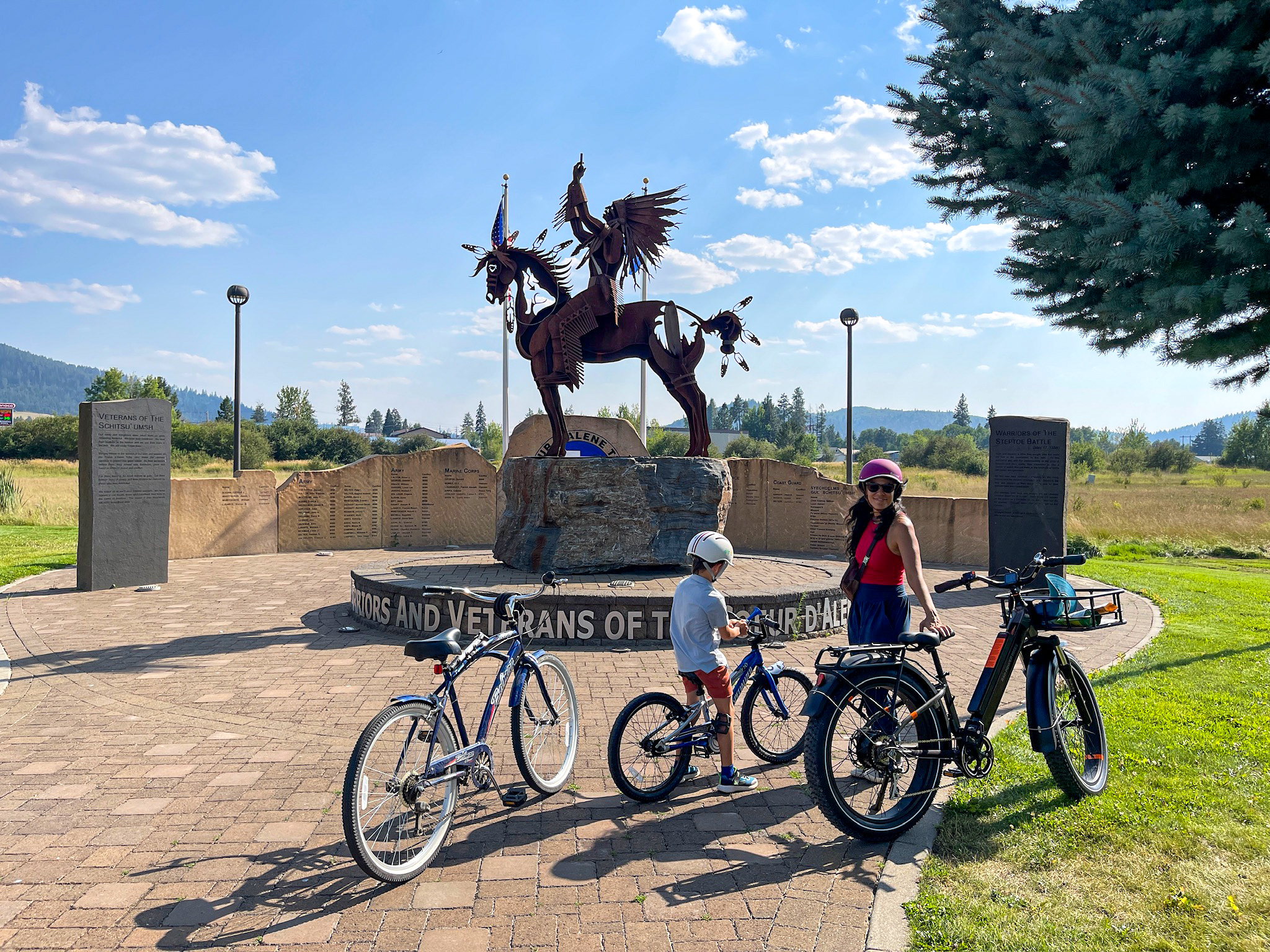 A woman and boy stand with their bikes on brick pavers in front of a large metal sculpture of a Native American warrior on horseback, part of the Warriors and Veterans of the Coeur d'Alene memorial in Plummer, Idaho, with inscribed stone panels and open grassland behind them.