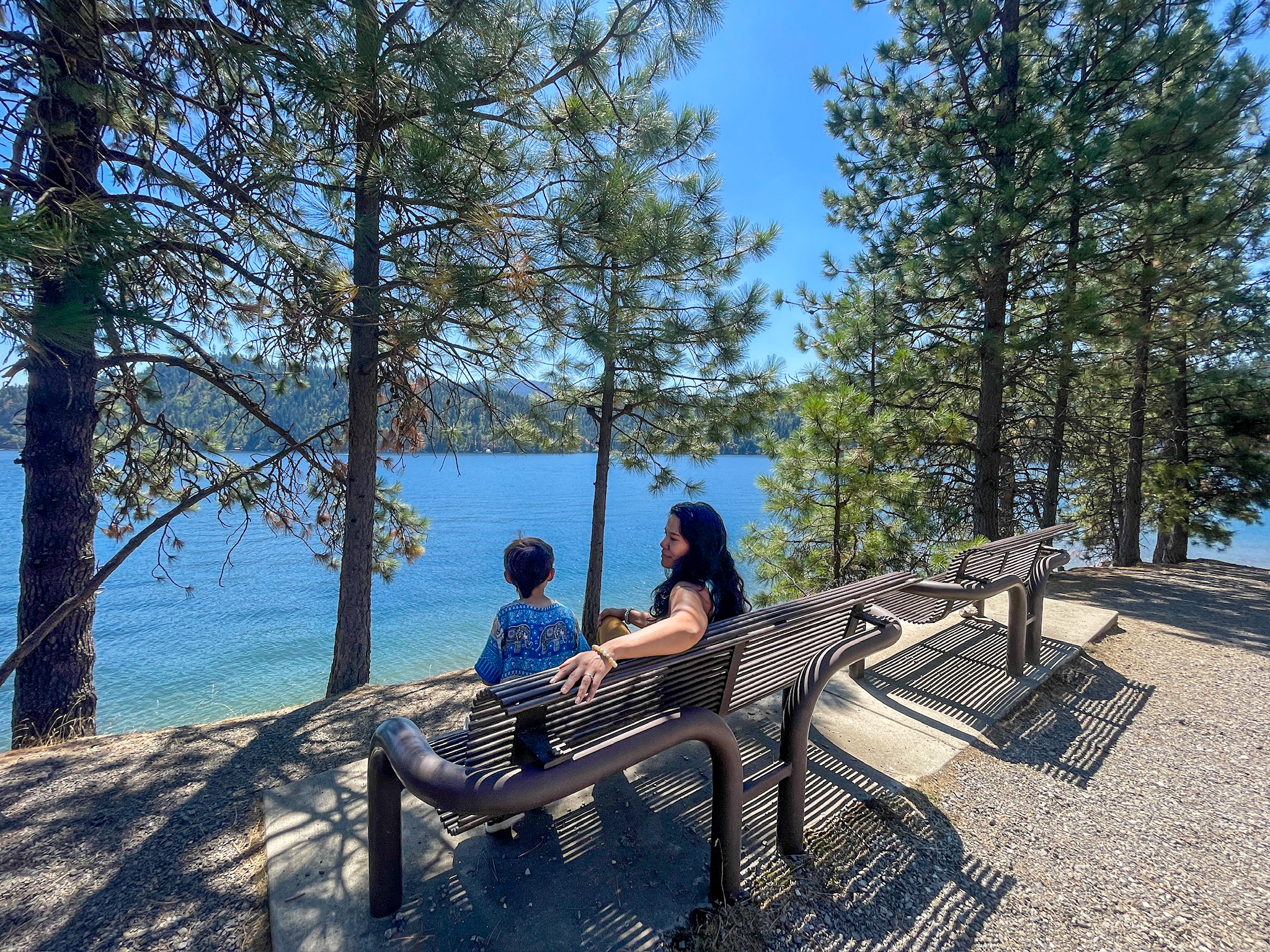 A woman and a young boy sit on a bench under tall pine trees, looking out over the deep blue water of Lake Coeur d'Alene near Higgens Point, with forested hills across the lake.