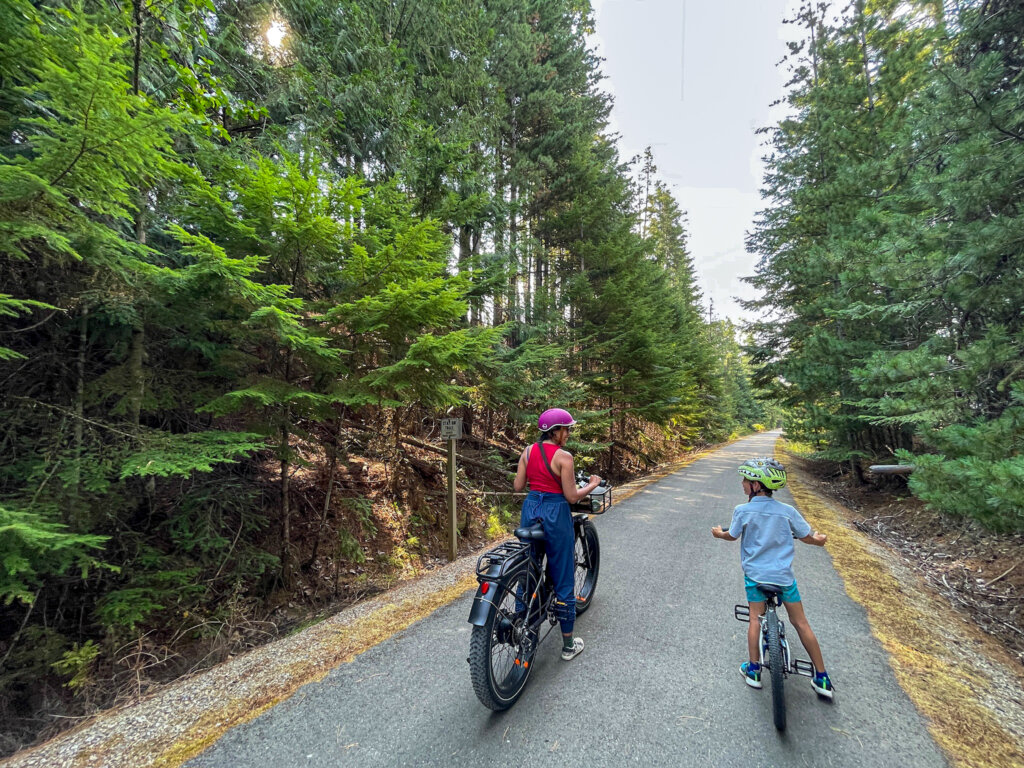 A woman and a boy on bicycles ride along the paved Trail of the Coeur d'Alenes trail lined with tall evergreen trees, with the path curving ahead into the forest.