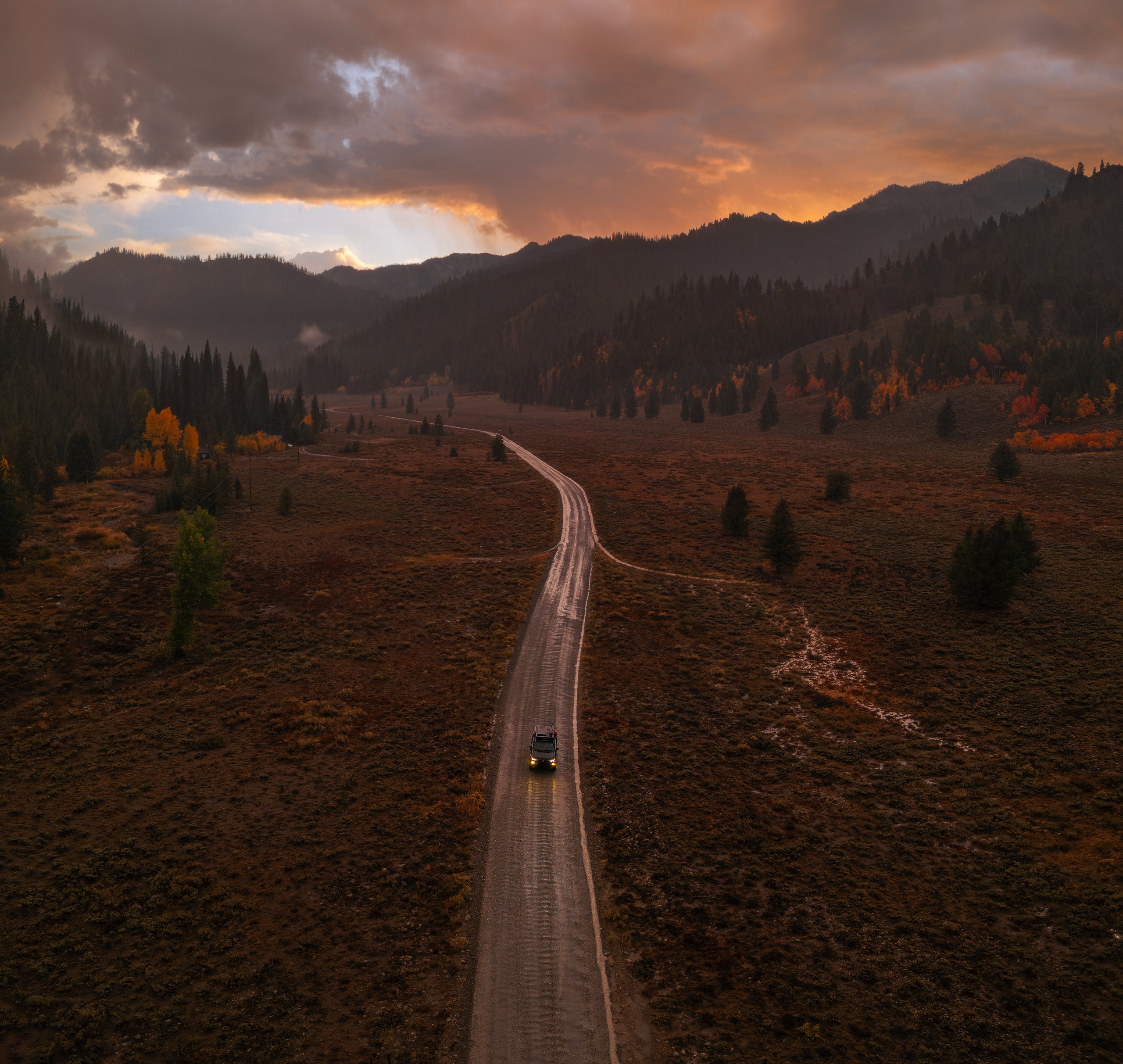 An aerial view of a vehicle with its headlights on, driving a wet gravel road through a wide sagebrush valley at sunset. The road winds back toward forested mountains, with patches of orange and yellow aspens on the slopes. Above, heavy storm clouds glow orange and pink, and light breaks through near the horizon.