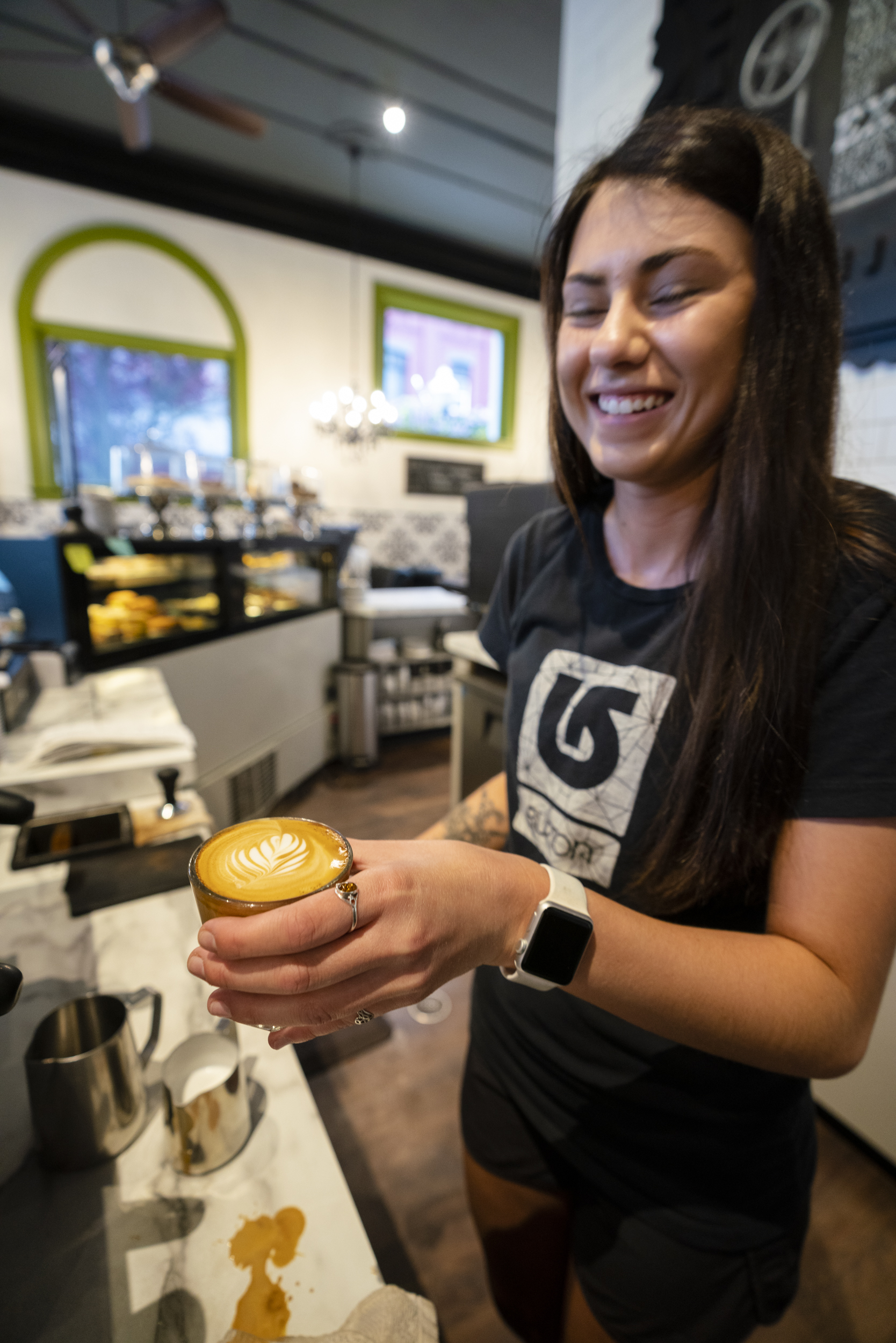 A smiling barista with long dark hair holds out a small glass cup of latte with rosetta foam art at The Vault counter.