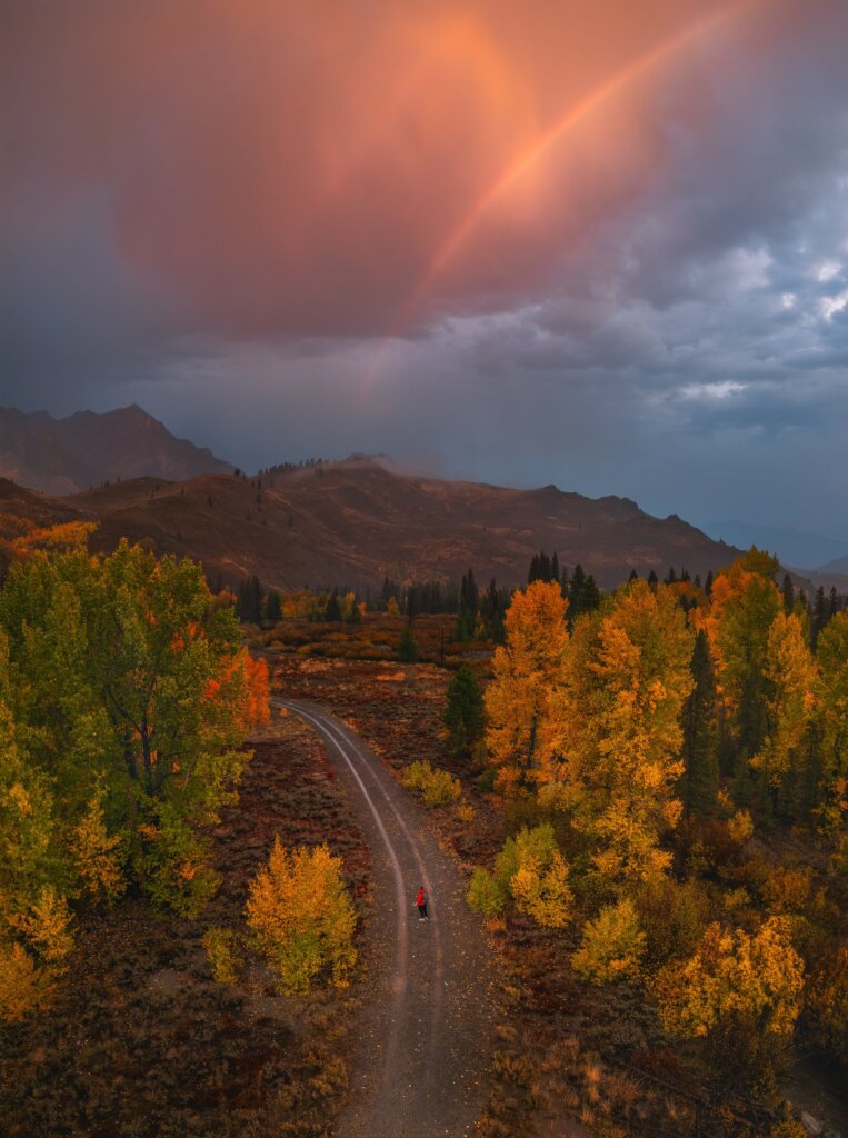 Aerial view of a hiker in a red jacket on the Harriman Trail near Ketchum, Idaho, among golden aspens under a rainbow.
