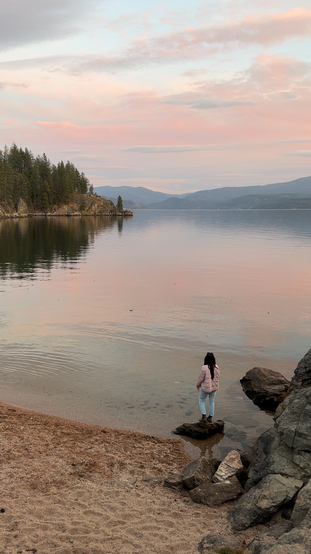 A person in a pink puffer jacket and light jeans stands on a rock at the edge of a calm lake at sunset, looking toward forested hills under a pink and blue sky.