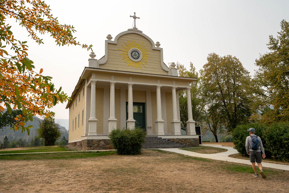 The Cataldo Mission near Coeur d’Alene, Idaho, a pale yellow wooden church with a columned front porch and a cross on its curved peak. A cyclist in a helmet with a blue backpack walks along a curving path toward the church. Dry grass, green trees and branches with autumn leaves frame the scene under a hazy sky.