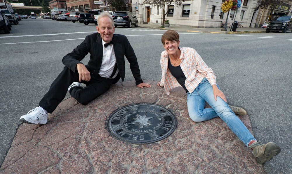 A smiling man in a black tuxedo jacket, bow tie and white sneakers sits on a red granite marker in the middle of a street in downtown Wallace, Idaho, next to a smiling woman in a plaid shirt and jeans. Between them is a round metal manhole cover with a compass star that declares Wallace the Center of the Universe. Brick and stone buildings, parked trucks and hanging flower baskets line the street behind them.